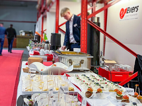 Mehrere Sorten Häppchen in Reihe auf einem langen Tisch, im Hintergrund ein Messestand und ein Mann im Anzug