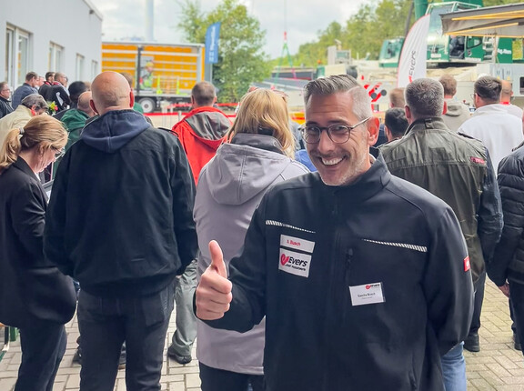 Ein lächelnder Mann mit Daumen-Hoch-Geste, im Hintergrund eine Gruppe an Menschen, die einer Vorführung zuschauen